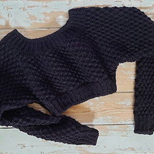 May include: A black knitted sweater with a textured pattern. The sweater has a wide neckline and long sleeves.