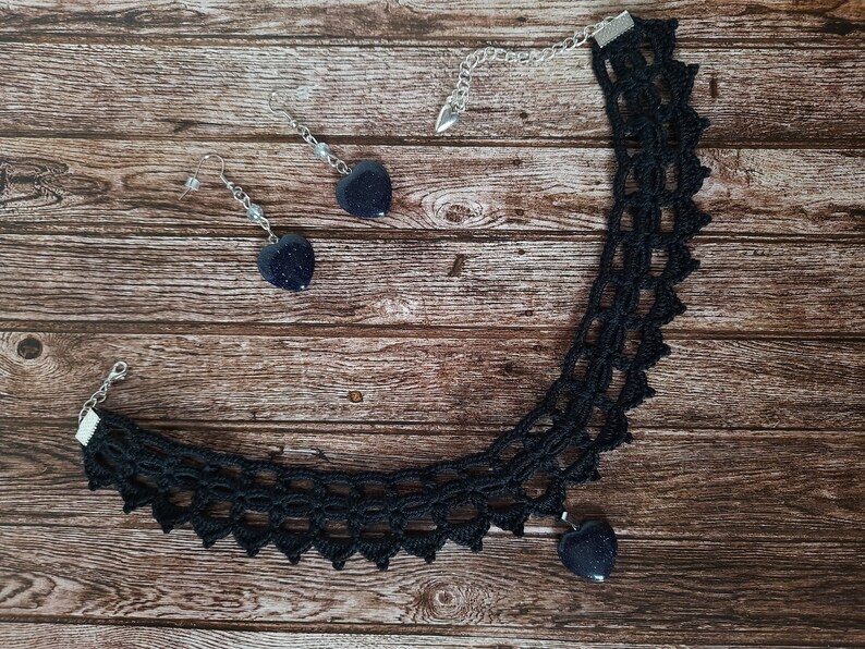 May include: A black crocheted choker necklace with a heart-shaped pendant and matching earrings. The pendant and earrings are made of dark blue stone.