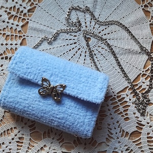 Puede incluir: Un bolso de peluche azul claro con un cierre de mariposa plateado y una correa de cadena plateada.