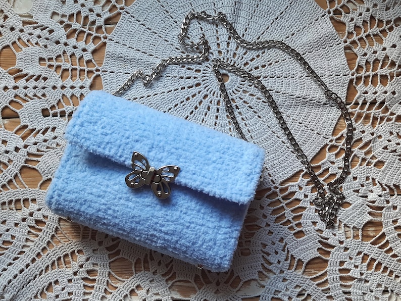 Puede incluir: Un bolso de peluche azul claro con un cierre de mariposa plateado y una correa de cadena plateada.