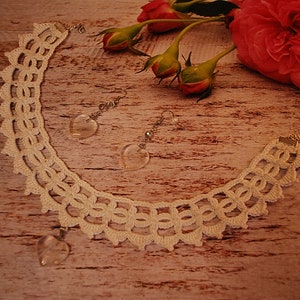 May include: A white crocheted necklace with a heart-shaped crystal pendant and matching earrings with heart-shaped crystal drops. The necklace and earrings are displayed on a wooden surface with pink roses.
