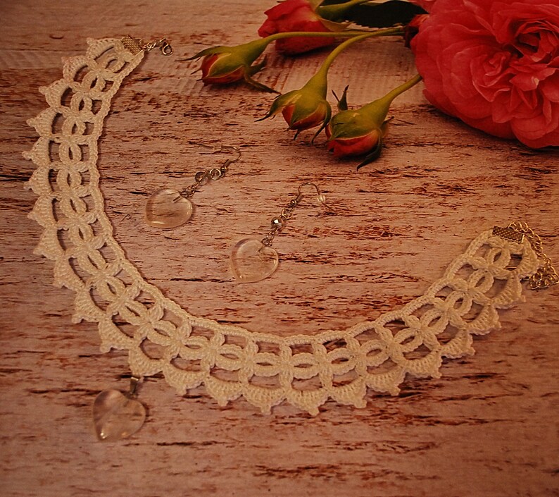 May include: A white crocheted necklace with a heart-shaped crystal pendant and matching earrings with heart-shaped crystal drops. The necklace and earrings are displayed on a wooden surface with pink roses.