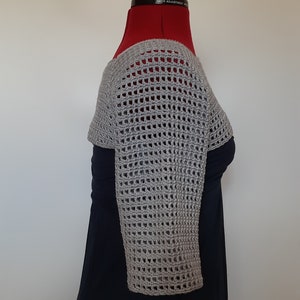 K&ouml;nnte beinhalten: Ein hellgrauer, offen gewebter H&auml;kel-Shrug mit langen &Auml;rmeln. Das Oberteil im Bolero-Stil wird auf einer roten Schneiderpuppe vor wei&szlig;em Hintergrund pr&auml;sentiert. Das H&auml;kelmuster weist rechteckige &Ouml;ffnungen auf, die ein zartes, luftiges Design erzeugen.