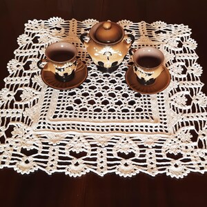 May include: White crocheted doily with a delicate floral pattern, featuring a brown teapot and two cups with saucers.