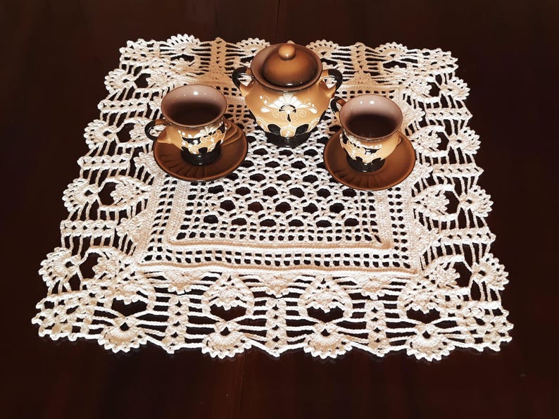 May include: White crocheted doily with a delicate floral pattern, featuring a brown teapot and two cups with saucers.