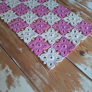 May include: A crocheted table runner featuring a pink and white floral design. The runner is crafted from fine, lacy stitches and is ideal for adding a touch of elegance to any table.