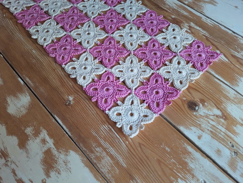 May include: A crocheted table runner featuring a pink and white floral design. The runner is crafted from fine, lacy stitches and is ideal for adding a touch of elegance to any table.