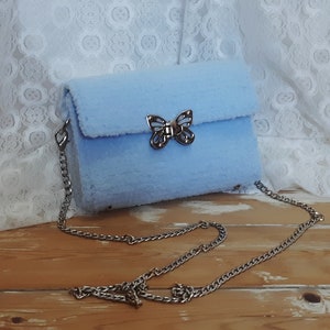 Puede incluir: Un bolso de hombro de color azul claro y peludo con un cierre de mariposa plateado y una correa de cadena plateada.
