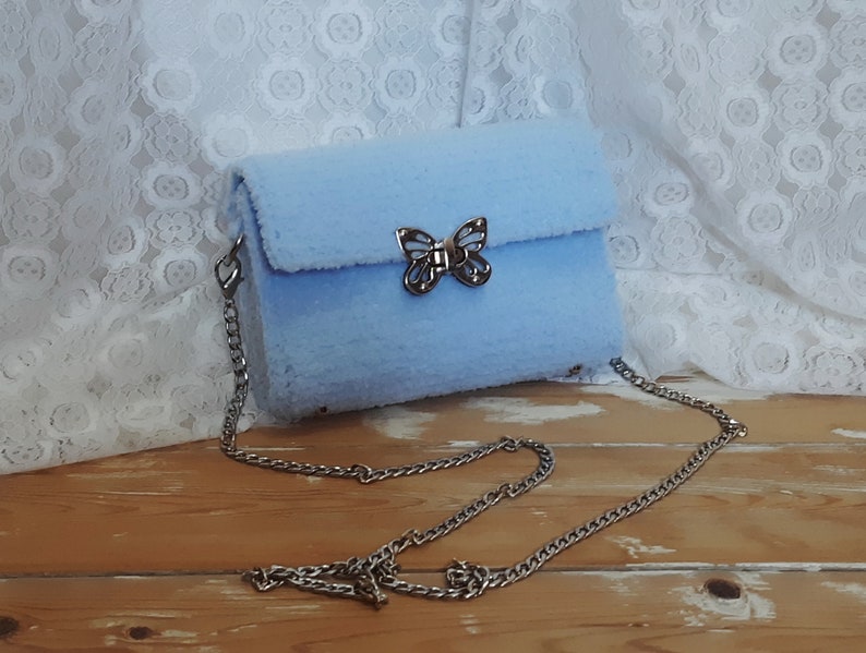 Puede incluir: Un bolso de hombro de color azul claro y peludo con un cierre de mariposa plateado y una correa de cadena plateada.