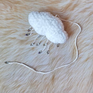 Puede incluir: Un art&iacute;culo de ganchillo blanco en forma de nube con cuentas iridiscentes colgantes. La nube tiene una textura esponjosa y est&aacute; conectada a un cord&oacute;n blanco. Las cuentas tienen forma de l&aacute;grima y reflejan varios colores. El art&iacute;culo es probablemente decorativo.