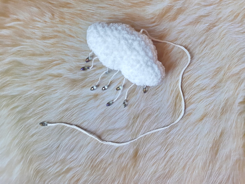 Puede incluir: Un art&iacute;culo de ganchillo blanco en forma de nube con cuentas iridiscentes colgantes. La nube tiene una textura esponjosa y est&aacute; conectada a un cord&oacute;n blanco. Las cuentas tienen forma de l&aacute;grima y reflejan varios colores. El art&iacute;culo es probablemente decorativo.