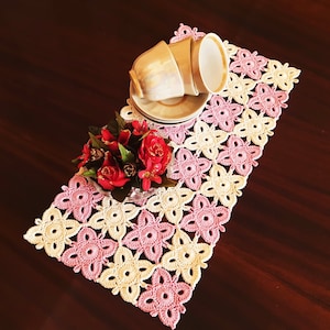 May include: A pink and cream crocheted table runner with a floral design. Two teacups and saucers are placed on the runner, and a bouquet of red roses is in the foreground.