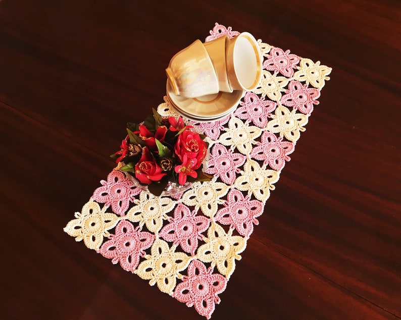 May include: A pink and cream crocheted table runner with a floral design. Two teacups and saucers are placed on the runner, and a bouquet of red roses is in the foreground.