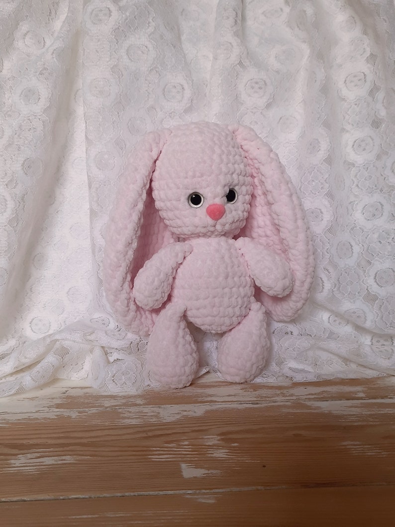 K&ouml;nnte beinhalten: Ein rosa geh&auml;keltes Hasenspielzeug mit schwarzen Augen und einer rosa Nase. Der Hase sitzt auf einem wei&szlig;en Spitzenstoff-Hintergrund.