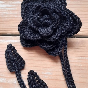 May include: A black crocheted flower with a stem made of three strands of yarn. The flower has multiple layers of petals and is a delicate and intricate design.