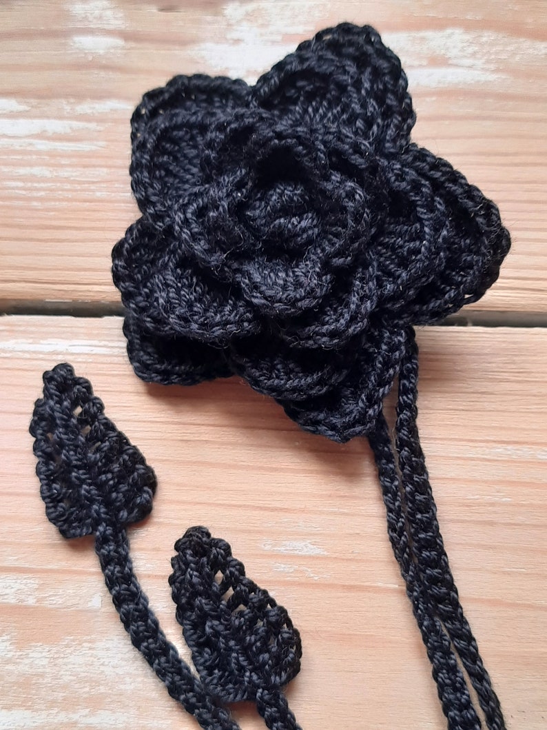 May include: A black crocheted flower with a stem made of three strands of yarn. The flower has multiple layers of petals and is a delicate and intricate design.