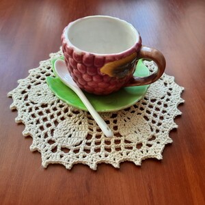 May include: A small, white ceramic teacup with a pink and brown raspberry design and a green leaf saucer. The cup and saucer are on a white crocheted doily.