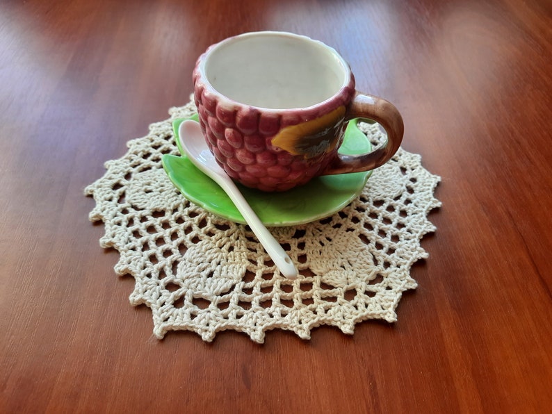 May include: A small, white ceramic teacup with a pink and brown raspberry design and a green leaf saucer. The cup and saucer are on a white crocheted doily.