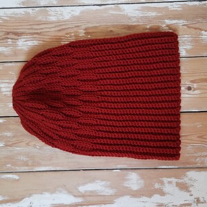 May include: A red knitted beanie hat with a ribbed texture. The hat is laid flat on a wooden surface.