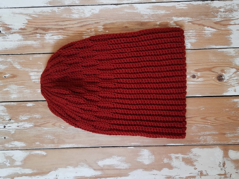May include: A red knitted beanie hat with a ribbed texture. The hat is laid flat on a wooden surface.