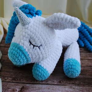 May include: A white crocheted unicorn with blue yarn accents. The unicorn has a silver horn and is lying down with its eyes closed.