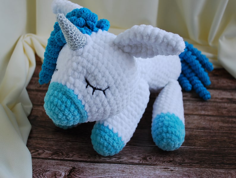 May include: A white crocheted unicorn with blue yarn accents. The unicorn has a silver horn and is lying down with its eyes closed.