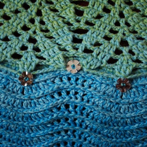 May include: A crocheted shawl with a green and blue ombre pattern. The shawl is decorated with silver flower-shaped embellishments.