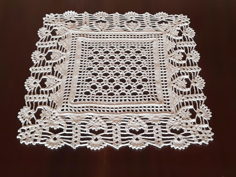 May include: White crocheted doily with a square center and a decorative border. The doily is made of delicate, intricate stitches.