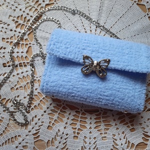 Puede incluir: Un bolso de peluche azul claro con un cierre de mariposa plateado y una correa de cadena plateada.