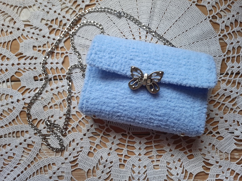 Puede incluir: Un bolso de peluche azul claro con un cierre de mariposa plateado y una correa de cadena plateada.