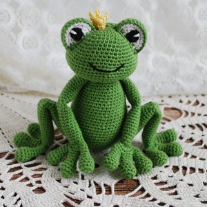 Pode incluir: Um sapo verde de croch&ecirc; feito &agrave; m&atilde;o com uma pequena coroa amarela. O sapo tem olhos grandes e redondos com detalhes brancos e pretos e est&aacute; sentado em posi&ccedil;&atilde;o vertical. As pernas do sapo est&atilde;o estendidas e ele est&aacute; colocado em uma superf&iacute;cie branca e rendada.