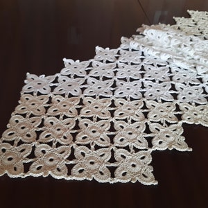 May include: White crocheted table runner with a delicate floral pattern. The runner is made of a fine thread and has a scalloped edge.