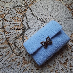 Puede incluir: Un bolso de peluche azul claro con un cierre de mariposa plateado y una correa de cadena plateada. El bolso est&aacute; sobre un encaje blanco de ganchillo.