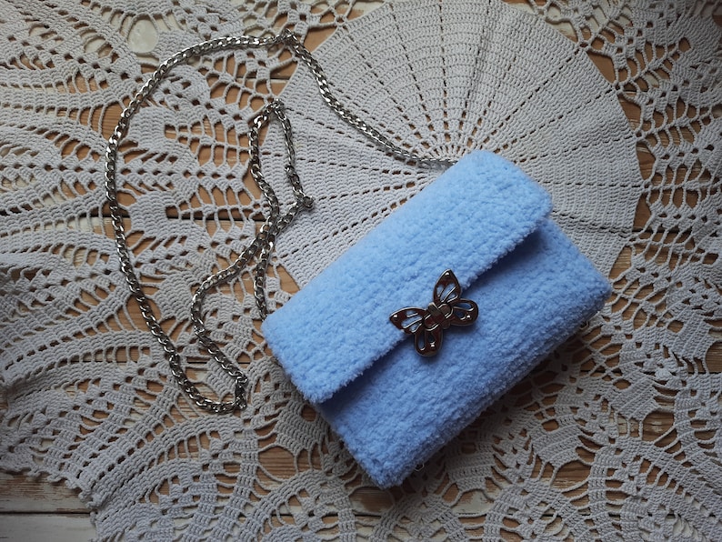 Puede incluir: Un bolso de peluche azul claro con un cierre de mariposa plateado y una correa de cadena plateada. El bolso est&aacute; sobre un encaje blanco de ganchillo.
