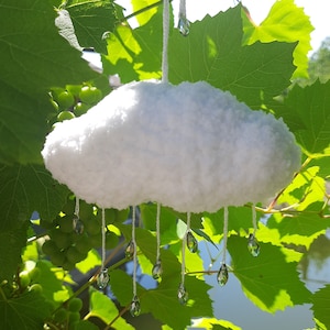 Puede incluir: M&oacute;vil en forma de nube blanca y esponjosa con l&aacute;grimas de cristal transparente colgando. La nube est&aacute; suspendida por un hilo blanco, sobre un fondo de hojas verdes de vid y luz solar.