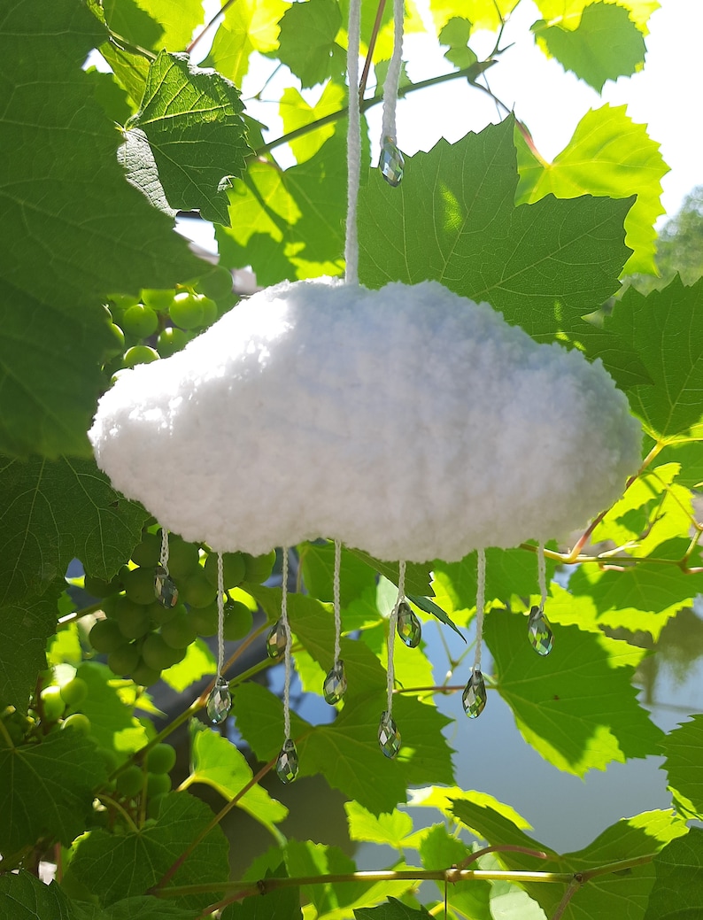 Puede incluir: M&oacute;vil en forma de nube blanca y esponjosa con l&aacute;grimas de cristal transparente colgando. La nube est&aacute; suspendida por un hilo blanco, sobre un fondo de hojas verdes de vid y luz solar.