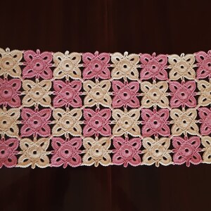 May include: A crocheted table runner with a pink and cream colour pattern. The runner is made of a delicate, lacy yarn and features a repeating floral design.
