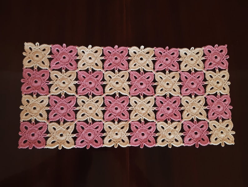 May include: A crocheted table runner with a pink and cream colour pattern. The runner is made of a delicate, lacy yarn and features a repeating floral design.