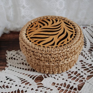 Pu&ograve; includere: Una scatola di immagazzinaggio rotonda in crochet con un coperchio in legno che presenta un design geometrico intagliato. La scatola &egrave; beige e il coperchio &egrave; di colore marrone chiaro.