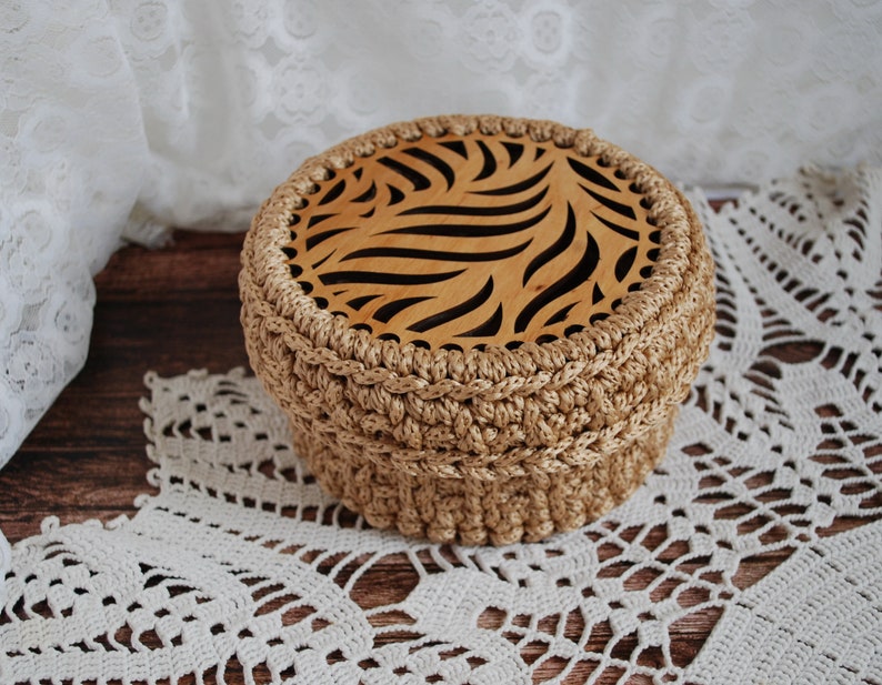 Pu&ograve; includere: Una scatola di immagazzinaggio rotonda in crochet con un coperchio in legno che presenta un design geometrico intagliato. La scatola &egrave; beige e il coperchio &egrave; di colore marrone chiaro.