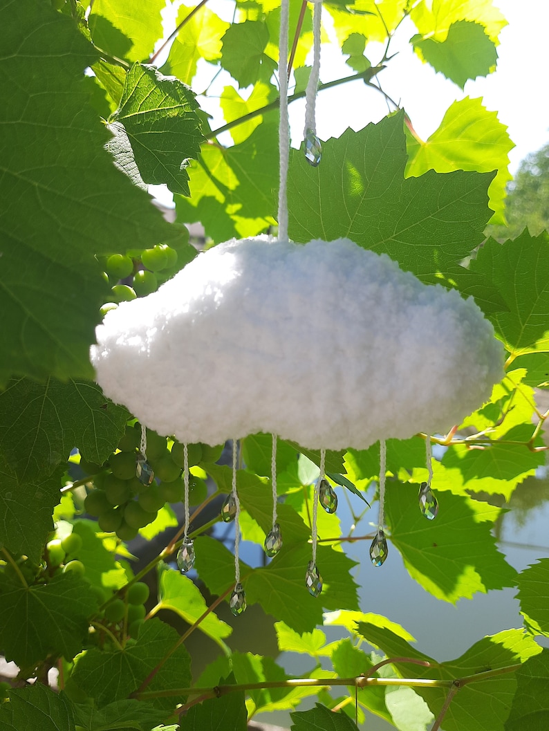 Puede incluir: Adorno en forma de nube blanca y esponjosa con l&aacute;grimas de cristal colgantes. La nube est&aacute; suspendida por una cuerda blanca, sobre un fondo de hojas verdes de vid y luz solar. Un art&iacute;culo decorativo caprichoso.