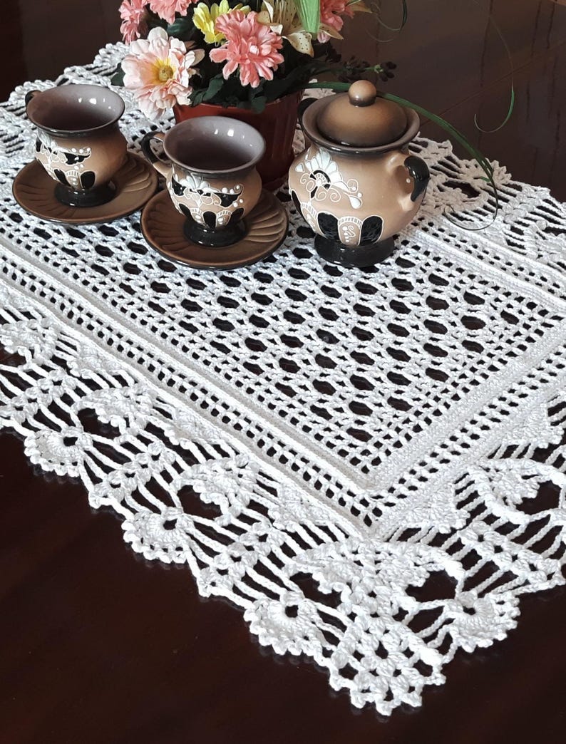 K&ouml;nnte beinhalten: Ein wei&szlig;er geh&auml;kelter Tischl&auml;ufer mit einem zarten Spitzenmuster. Der L&auml;ufer liegt auf einem dunklen Holztisch und zeigt ein Set aus drei Keramik-Teetassen und einer Zuckerdose mit Blumendesigns.