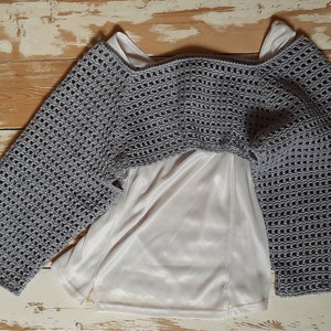 K&ouml;nnte beinhalten: Ein graues H&auml;kel-Bolero mit langen &Auml;rmeln, getragen &uuml;ber einem wei&szlig;en Satin-Top. Das Bolero hat ein offenes, luftiges Gewebe. Die &Auml;rmel sind weit und ausgestellt. Das Kleidungsst&uuml;ck wird auf einer hellen Holzoberfl&auml;che pr&auml;sentiert.