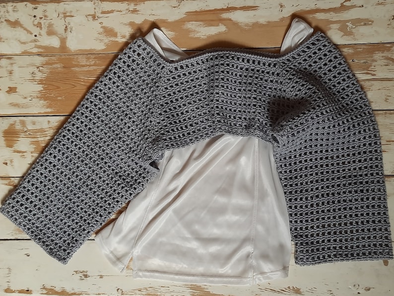 K&ouml;nnte beinhalten: Ein graues H&auml;kel-Bolero mit langen &Auml;rmeln, getragen &uuml;ber einem wei&szlig;en Satin-Top. Das Bolero hat ein offenes, luftiges Gewebe. Die &Auml;rmel sind weit und ausgestellt. Das Kleidungsst&uuml;ck wird auf einer hellen Holzoberfl&auml;che pr&auml;sentiert.