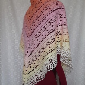 May include: A handmade, crocheted shawl in a gradient of colors, from peach to pink to yellow, with a white lace trim. The shawl is draped over a mannequin, showcasing its intricate openwork design and soft color palette.