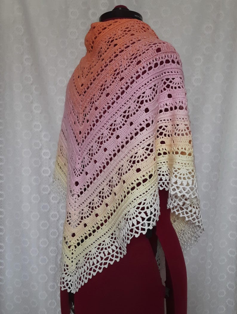 May include: A handmade, crocheted shawl in a gradient of colors, from peach to pink to yellow, with a white lace trim. The shawl is draped over a mannequin, showcasing its intricate openwork design and soft color palette.