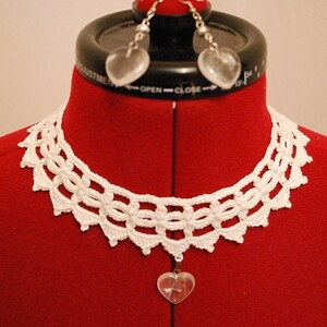 May include: White crocheted choker necklace with a heart-shaped crystal pendant. The necklace is on a red fabric mannequin. The mannequin is also wearing a pair of heart-shaped crystal earrings.