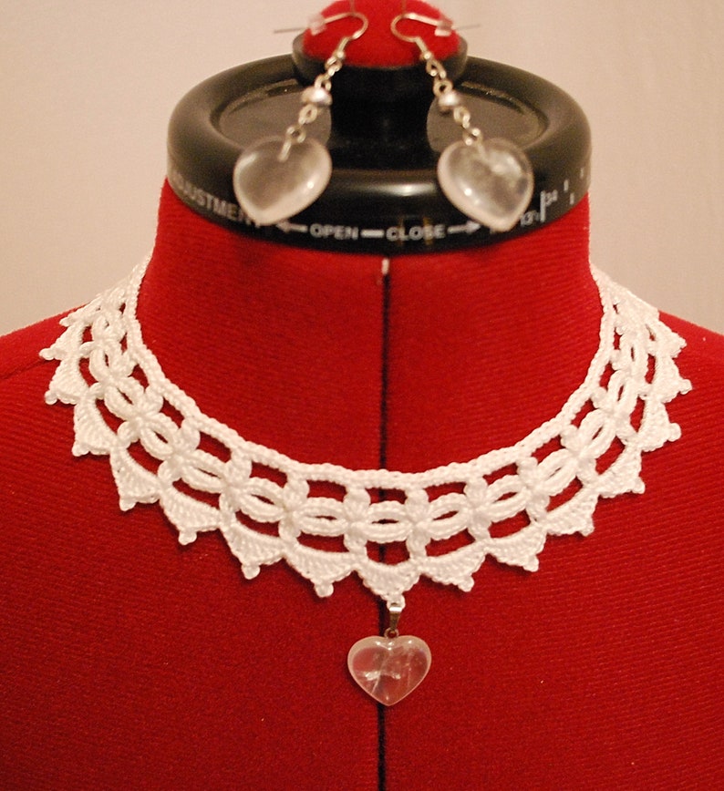 May include: White crocheted choker necklace with a heart-shaped crystal pendant. The necklace is on a red fabric mannequin. The mannequin is also wearing a pair of heart-shaped crystal earrings.