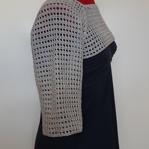 K&ouml;nnte beinhalten: Ein hellgrauer, offen gewebter H&auml;kel-Bolero mit Dreiviertel&auml;rmeln. Der Bolero wird auf einer Schaufensterpuppe &uuml;ber einem schwarzen Kleid pr&auml;sentiert. Das H&auml;kelmuster weist ein gitterartiges Design auf.