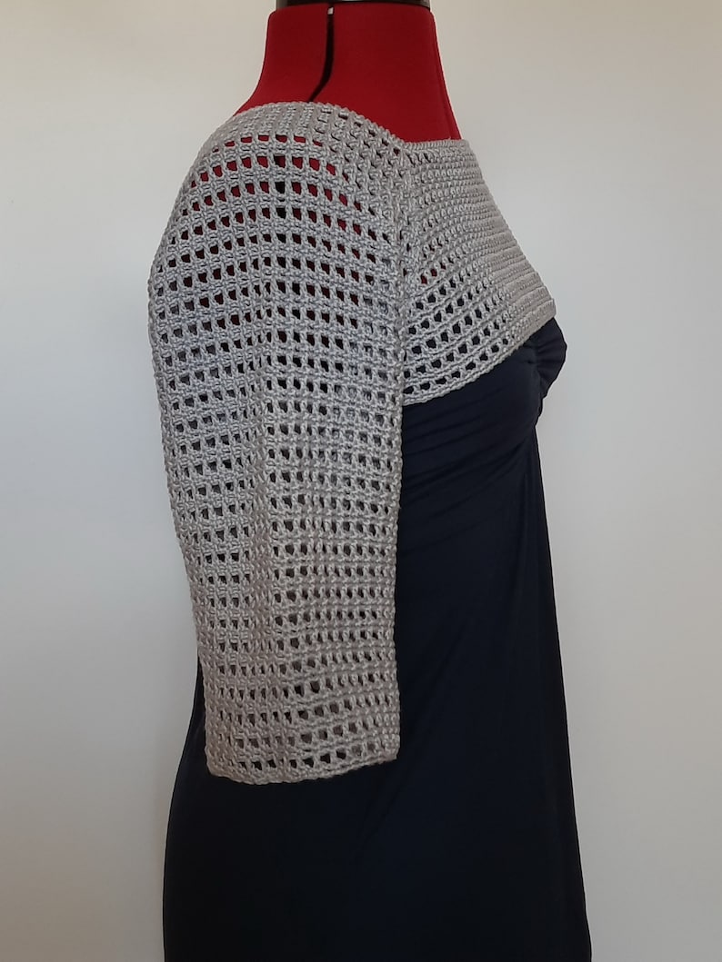 K&ouml;nnte beinhalten: Ein hellgrauer, offen gewebter H&auml;kel-Bolero mit Dreiviertel&auml;rmeln. Der Bolero wird auf einer Schaufensterpuppe &uuml;ber einem schwarzen Kleid pr&auml;sentiert. Das H&auml;kelmuster weist ein gitterartiges Design auf.